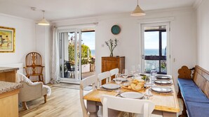 A dining area with a wooden table set for four, a bench, and a view of the sea through large glass doors. - Penrose - Beach House With Direct Beach Access (Saundersfoot)