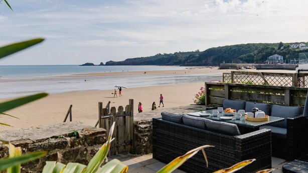 A beachside restaurant with outdoor seating overlooking the sea, a wooden fence, and a view of the beach and hills in the background. - Penrose - Beach House With Direct Beach Access (Saundersfoot)