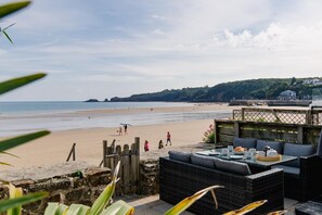 Un restaurant situé en bord de mer, avec une terrasse offrant une vue sur l’océan, une clôture en bois et une vue panoramique sur la plage et les collines en arrière-plan.