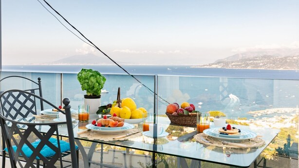 Un tavolo da balcone con prodotti per la colazione, una pianta in vaso e vista sul mare e sulle montagne.