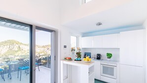 Una cucina moderna con mobili bianchi, forno integrato e una piccola isola con lavello. Si vede un balcone con sedie blu e vista su un paesaggio montano.
