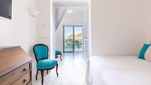 Camera d'albergo con un letto, una cassettiera in legno, una sedia blu e vista all'aperto tramite una porta scorrevole in vetro.