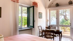 A bedroom with a bed, a table, chairs, and a view of greenery through the window. - Villa Ginestra by Elite Villas (Capri)