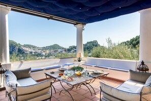 Une terrasse extérieure avec une table dressée pour le dîner, deux canapés et une vue sur une ville située sur une colline.