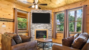 Een gezellig cabin interieur met een open haard, leren banken, een flatscreen televisie en grote ramen met uitzicht op het bos.