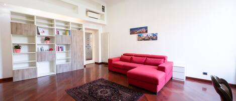 A spacious living room with a red sectional sofa, a large wooden floor, and a built-in white shelving unit with decorative items.