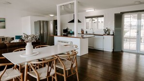 A modern kitchen with a central island, white cabinetry, and a dining area with a white table and wicker chairs. - THE BILLOWS - BINALONG BAY.  Hamptons style escape overlooking the Bay of Fires. (Binalong Bay)