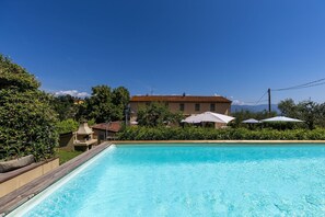 A swimming pool with a wooden deck, surrounded by greenery and a building with a tiled roof.