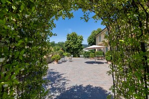 An outdoor patio area with tables and chairs, surrounded by greenery and a building in the background.