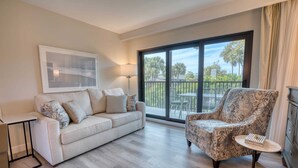 A living room with a sofa, armchair, and a table with a book. There is a large window with a view of trees and a balcony. - Newly Renovated Park Shore Resort Deluxe Suite 238 (Naples)