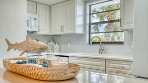 A kitchen with white cabinets, a microwave, a window with a view of palm trees, and a decorative fish sculpture on the counter. - Newly Renovated Park Shore Resort Deluxe Suite 238 (Naples)