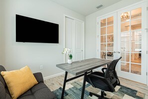 A modern living room with a flat-screen TV mounted on the wall, a dining table with chairs, a sofa with a cushion, and a large glass door leading to a dining area.