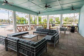 A covered outdoor seating area with sofas, tables, and umbrellas, overlooking a pool and greenery.