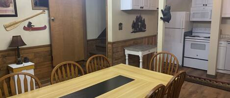 Un comedor con una mesa y sillas de madera, una mesita blanca con una lámpara, y una zona de cocina con armarios y electrodomésticos blancos.