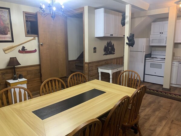 Un comedor con una mesa y sillas de madera, una mesita blanca con una lámpara, y una zona de cocina con armarios y electrodomésticos blancos.