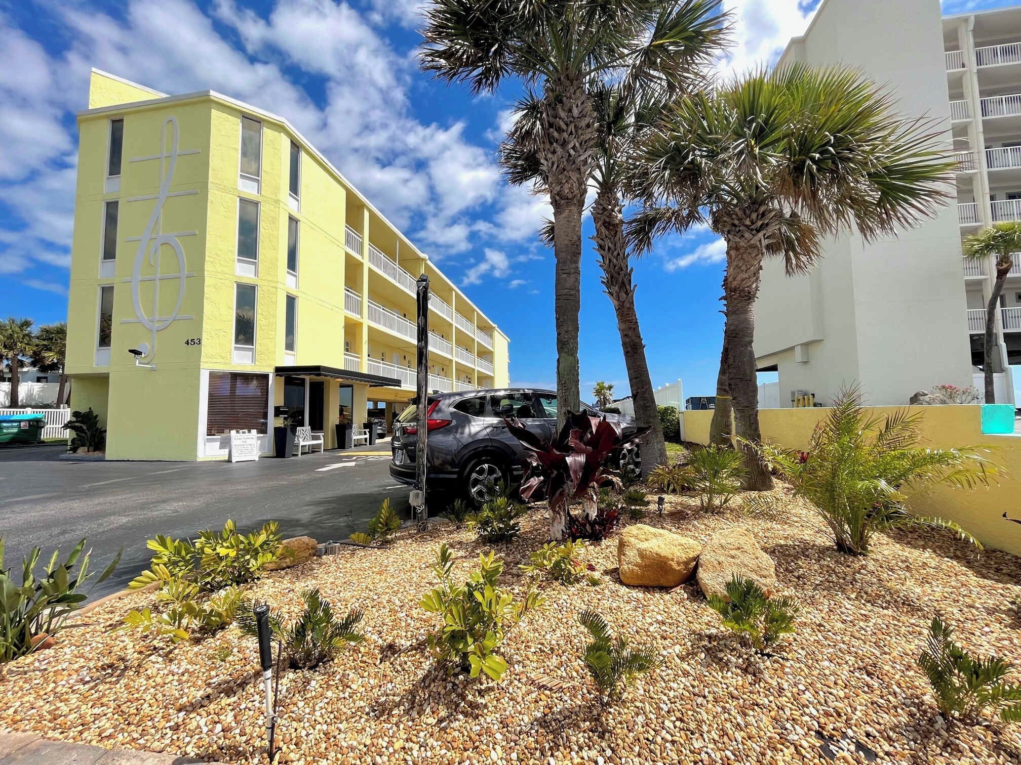 Un edificio amarillo con varios balcones, un área de estacionamiento con coches y un jardín ajardinado con palmeras y piedras.