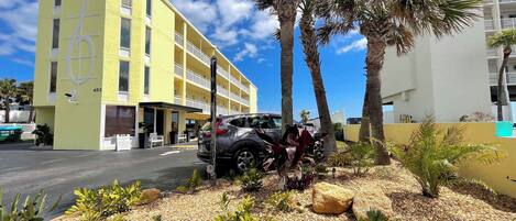 Un edificio amarillo con varios balcones, un área de estacionamiento con coches y un jardín ajardinado con palmeras y piedras.