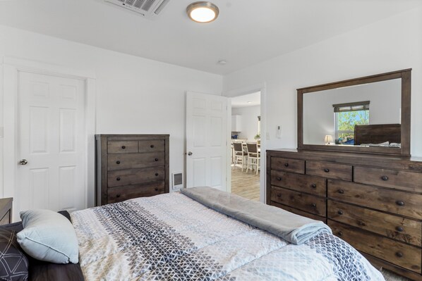A bedroom with a bed, a dresser, a mirror, and a door leading to a dining area.