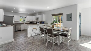 A modern kitchen with white cabinetry, a dining area with a table and chairs, and a large window providing natural light. - Amber Nook! Always clean, and build to be comfortable! (The Dalles)