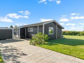 Une maison de plain-pied à l’extérieur de couleur gris foncé, avec un toit en tuiles et un petit jardin devant, composé d’une pelouse et d’un jeune arbre.
