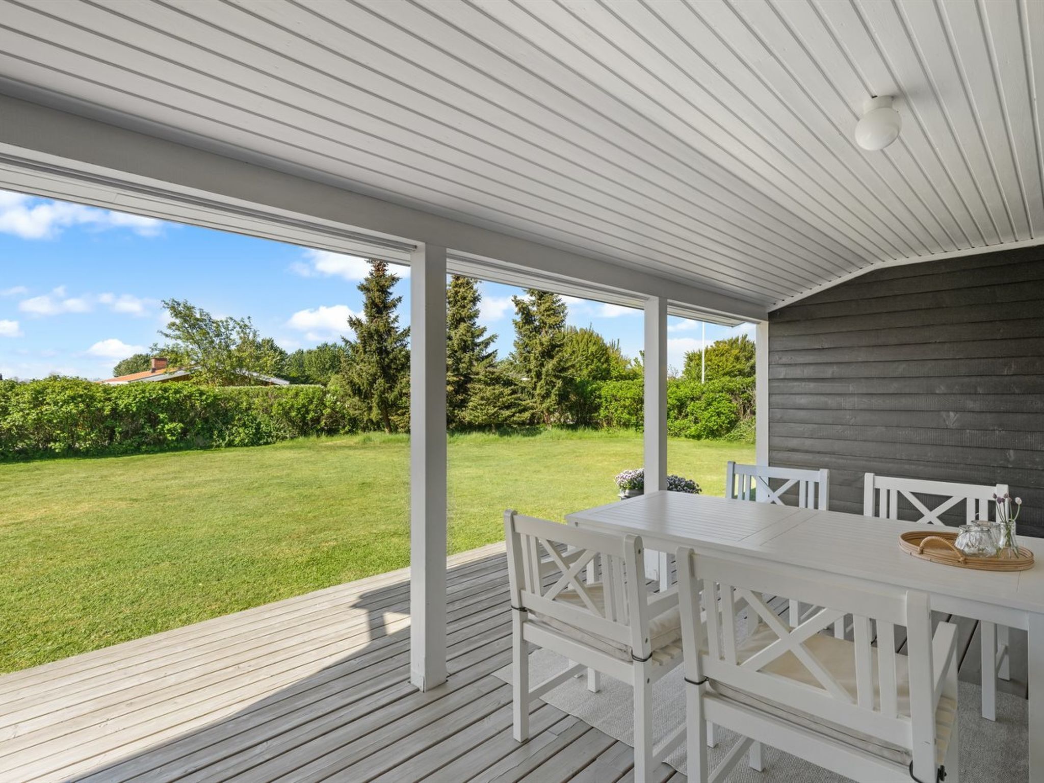A covered outdoor area with a white table and chairs, a wooden deck, and a view of a green lawn and trees.