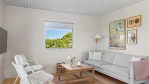 A modern living room with a white sofa, a wooden coffee table, and a white chair with a fluffy cushion. There is a window with a view of greenery outside. - "Iven" - 140m from the sea by Interhome (Otterup)