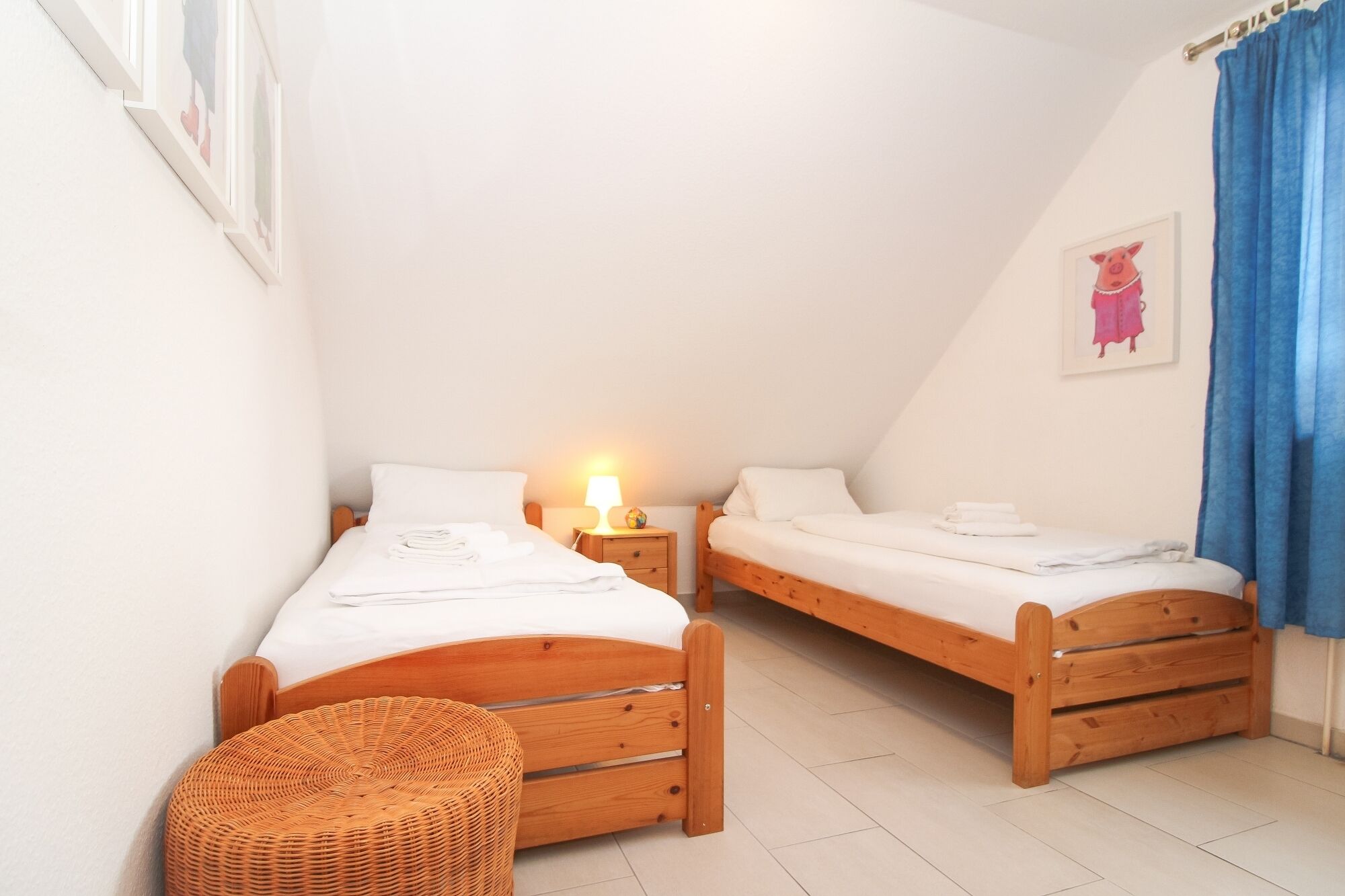 A small, cozy room with two wooden beds, a wicker chair, and a nightstand with a lamp.