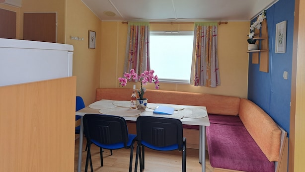 A compact room with a table, two chairs, a bench, a window with curtains, a clock, and a small shelf with decorative items. - Joli Mobil Home sur Camping (Enquin sur baillons)