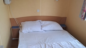 A single bed with white bedding, a wooden headboard, and a nightstand with a lamp. - Joli Mobil Home sur Camping (Enquin sur baillons)