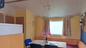 A compact room with a dining table, two blue chairs, a bench, and a window with curtains. - Joli Mobil Home sur Camping (Enquin sur baillons)