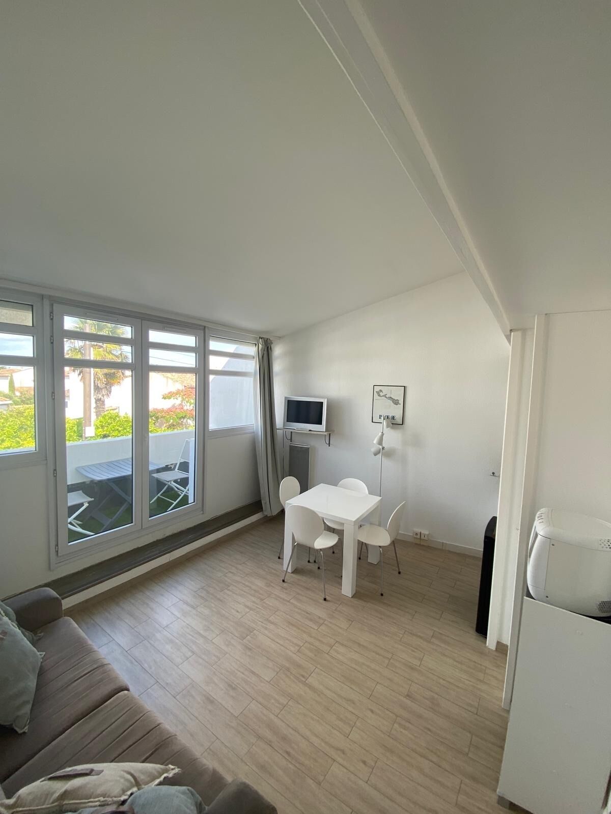 Una habitación pequeña y luminosa con una mesa y sillas blancas, un televisor y un refrigerador.
