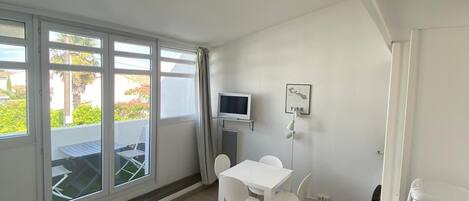 Una habitación pequeña y luminosa con una mesa y sillas blancas, un televisor y un refrigerador.