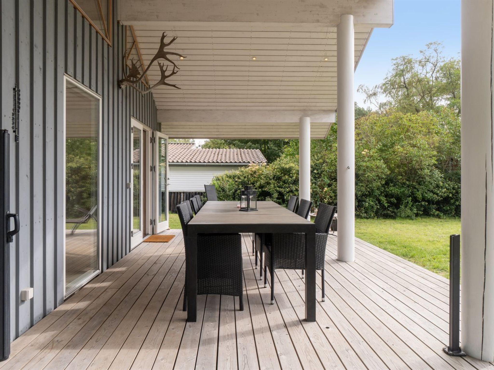 A covered patio with a wooden deck, a dining table with chairs, and a view of a house and greenery in the background.