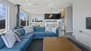 A modern living room with a blue sectional sofa, a wooden coffee table, and a kitchen area with white cabinets and stainless steel appliances. - Stylish Serenity - Martinborough Holiday Home (Martinborough)