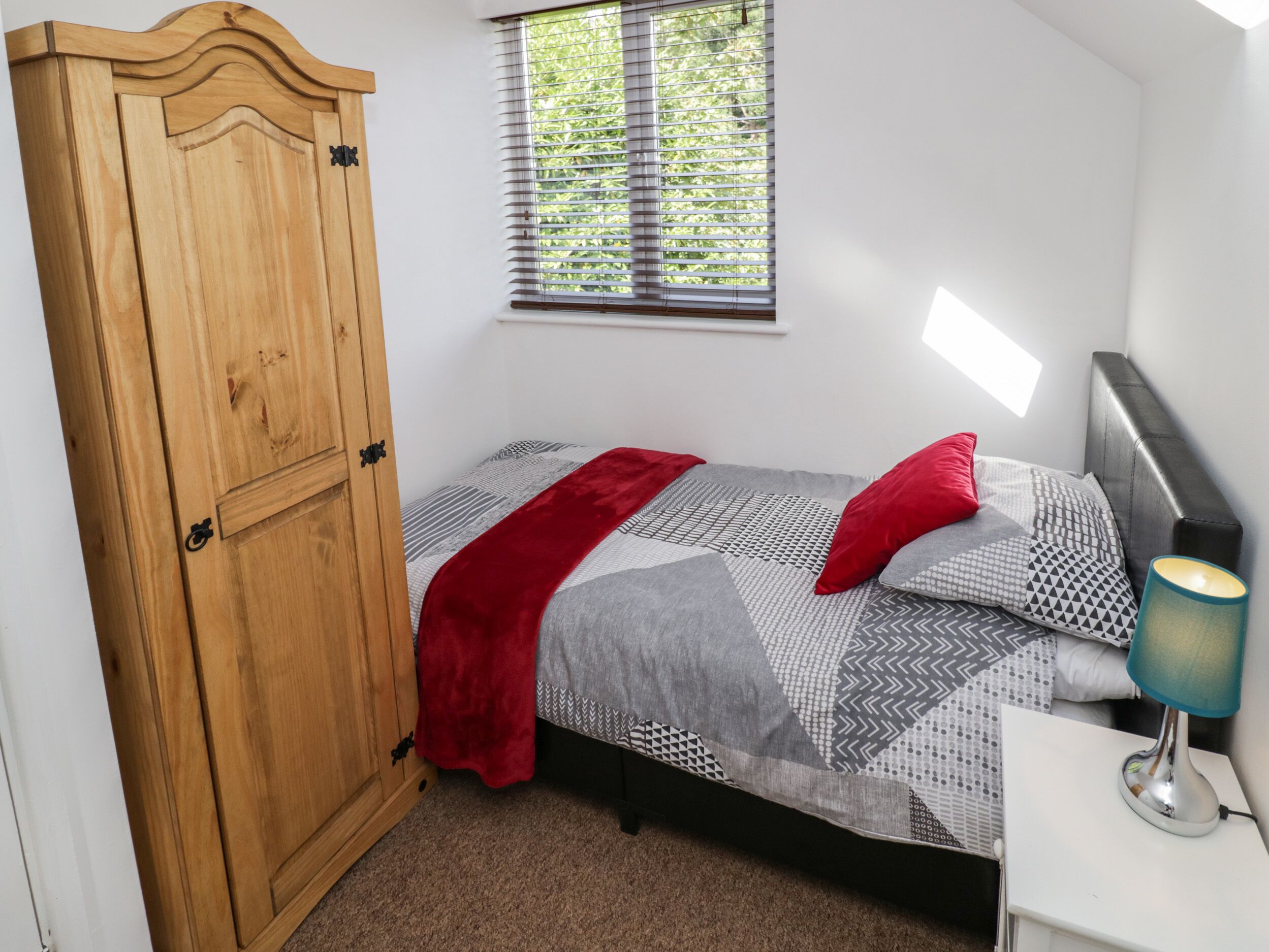 A bedroom with a bed, a wooden wardrobe, a window with blinds, and a bedside lamp.