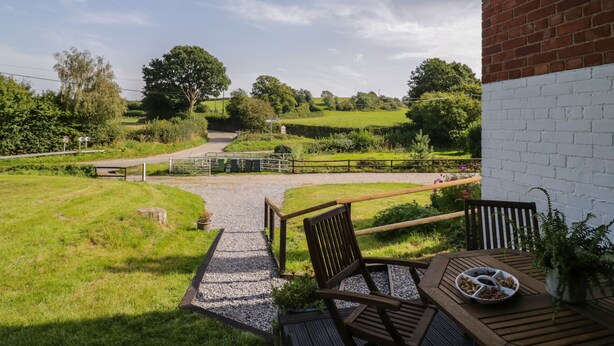 A patio with a table and chairs, a gravel path leading to a road, and a rural landscape with fields and trees. - 3 Oaks Lodge (Shrewsbury)