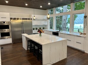 Une cuisine moderne dotée d’un grand îlot central, d’appareils électroménagers en acier inoxydable, de luminaires suspendus et de grandes fenêtres offrant une vue sur l’extérieur.