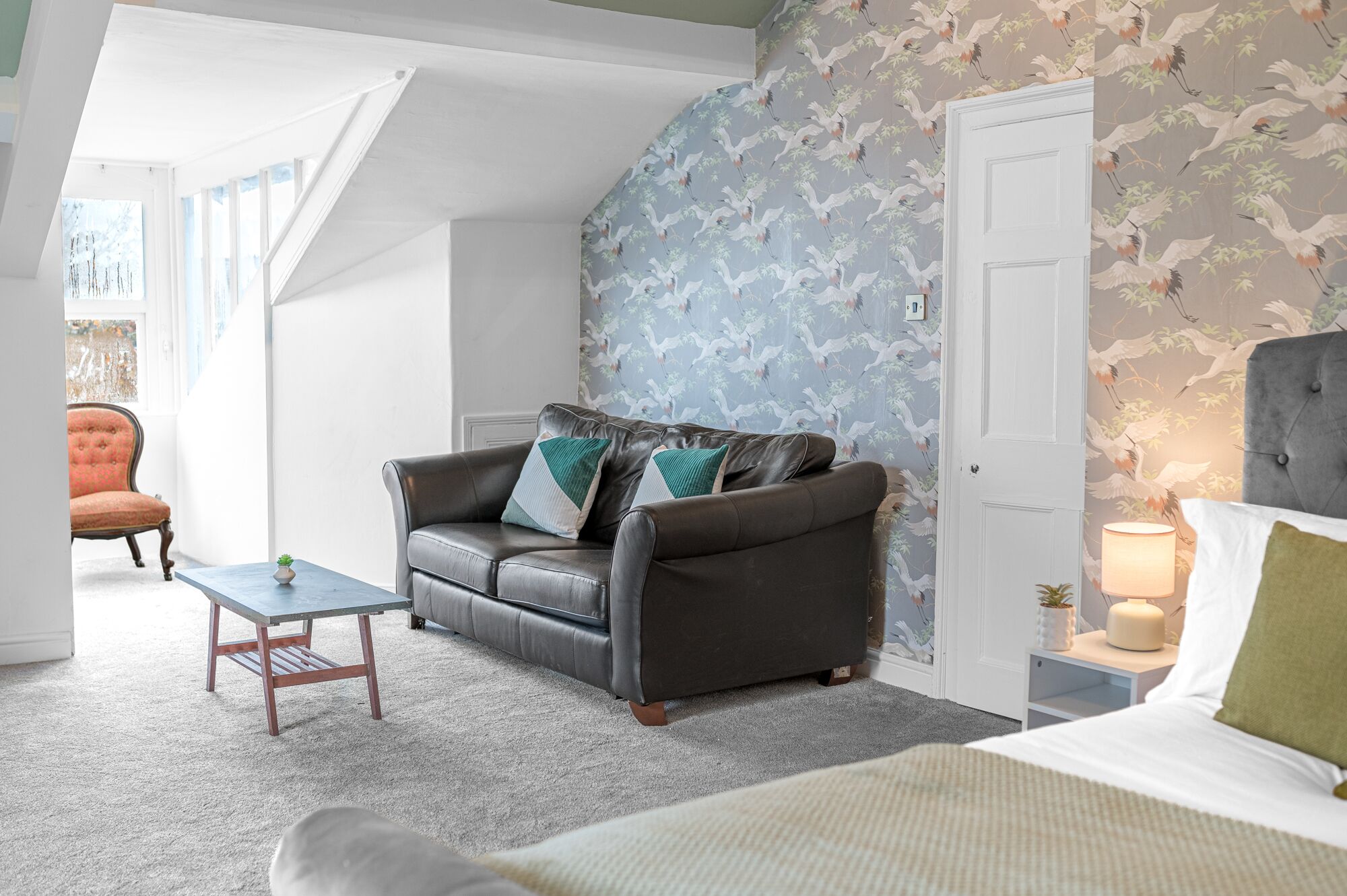Ein gemütlicher Raum mit einem Ledersofa, einem kleinen Tisch und einem Bett. Eine Tür mit floralem Wandpapier und ein Fenster mit Blick.