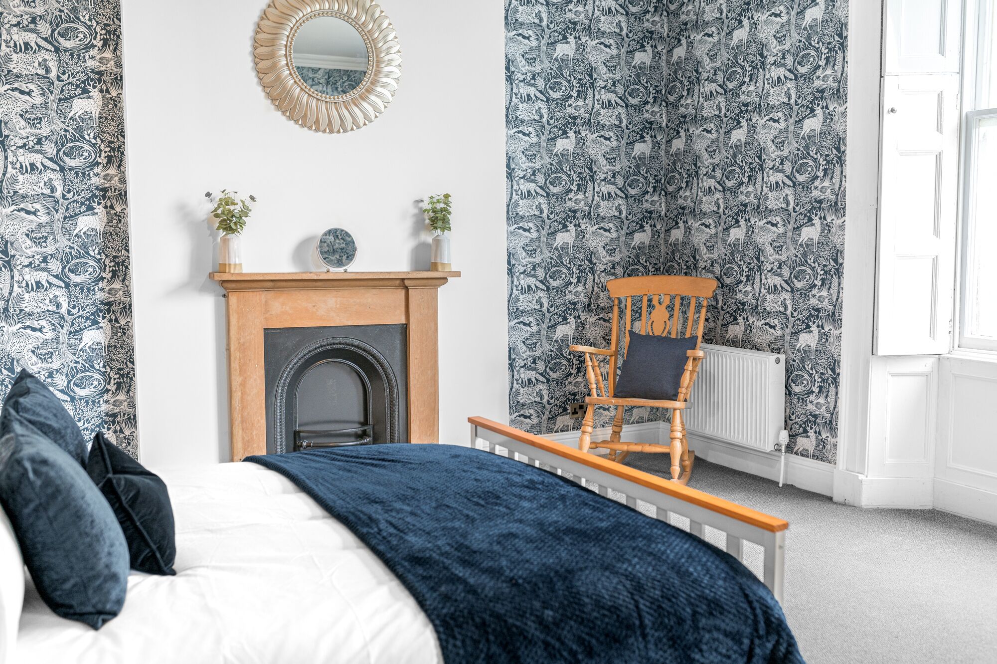 A bedroom with a bed, a wooden chair, a fireplace, and patterned wallpaper.