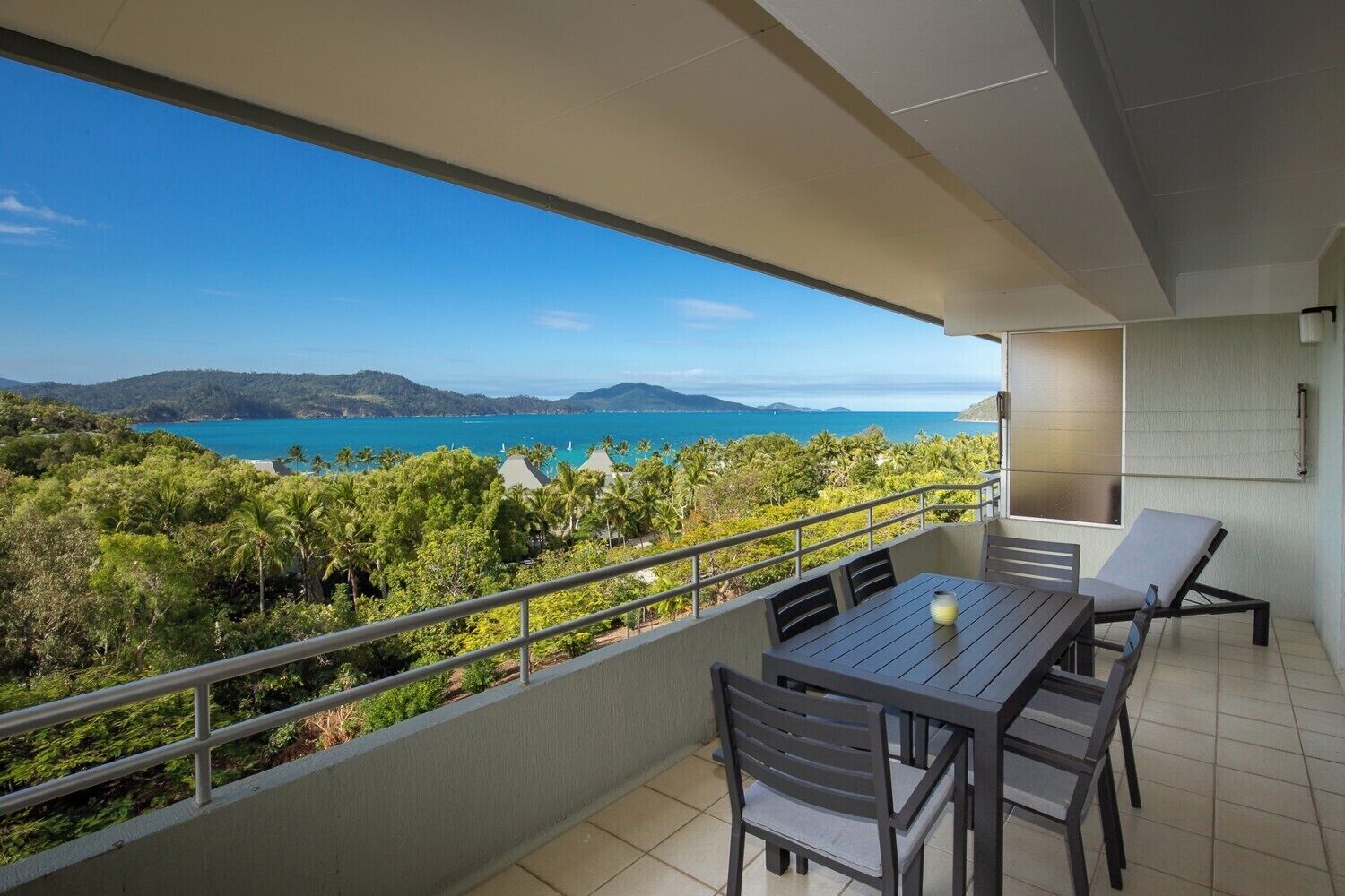 A balcony with a table and chairs, offering a view of the sea and hills.