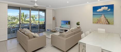 A modern living room with a glass dining table, two sofas, a TV, and a large window with a view of palm trees and a beach.