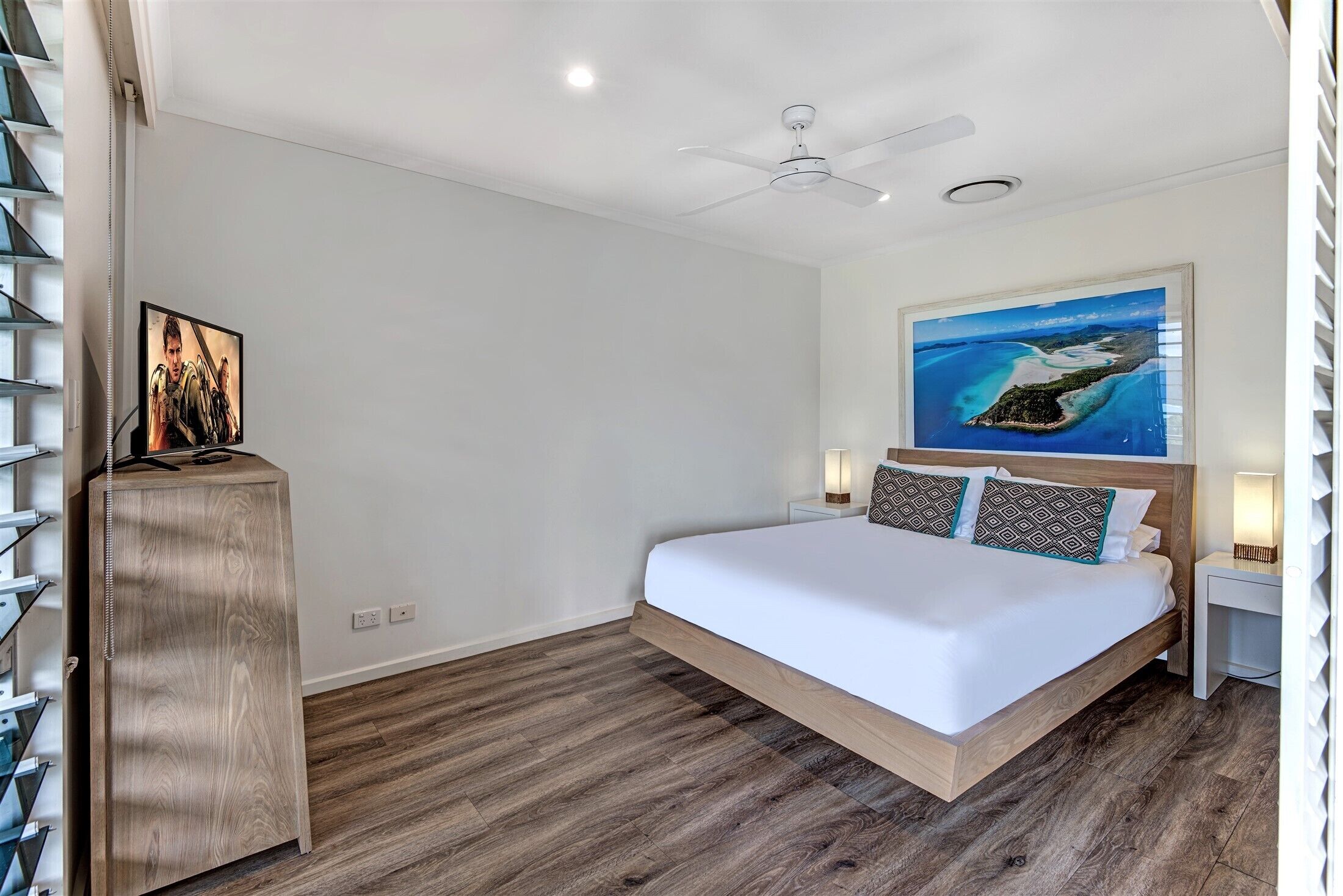 A bedroom with a large bed, a ceiling fan, a painting of a tropical island, and a wooden nightstand.