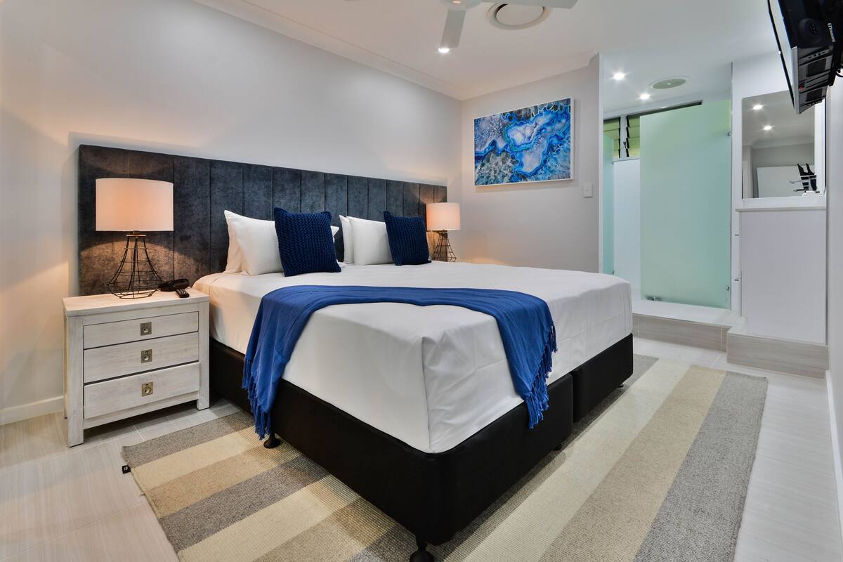 A modern bedroom with a large bed, two bedside tables, a blue throw blanket, and a painting on the wall.