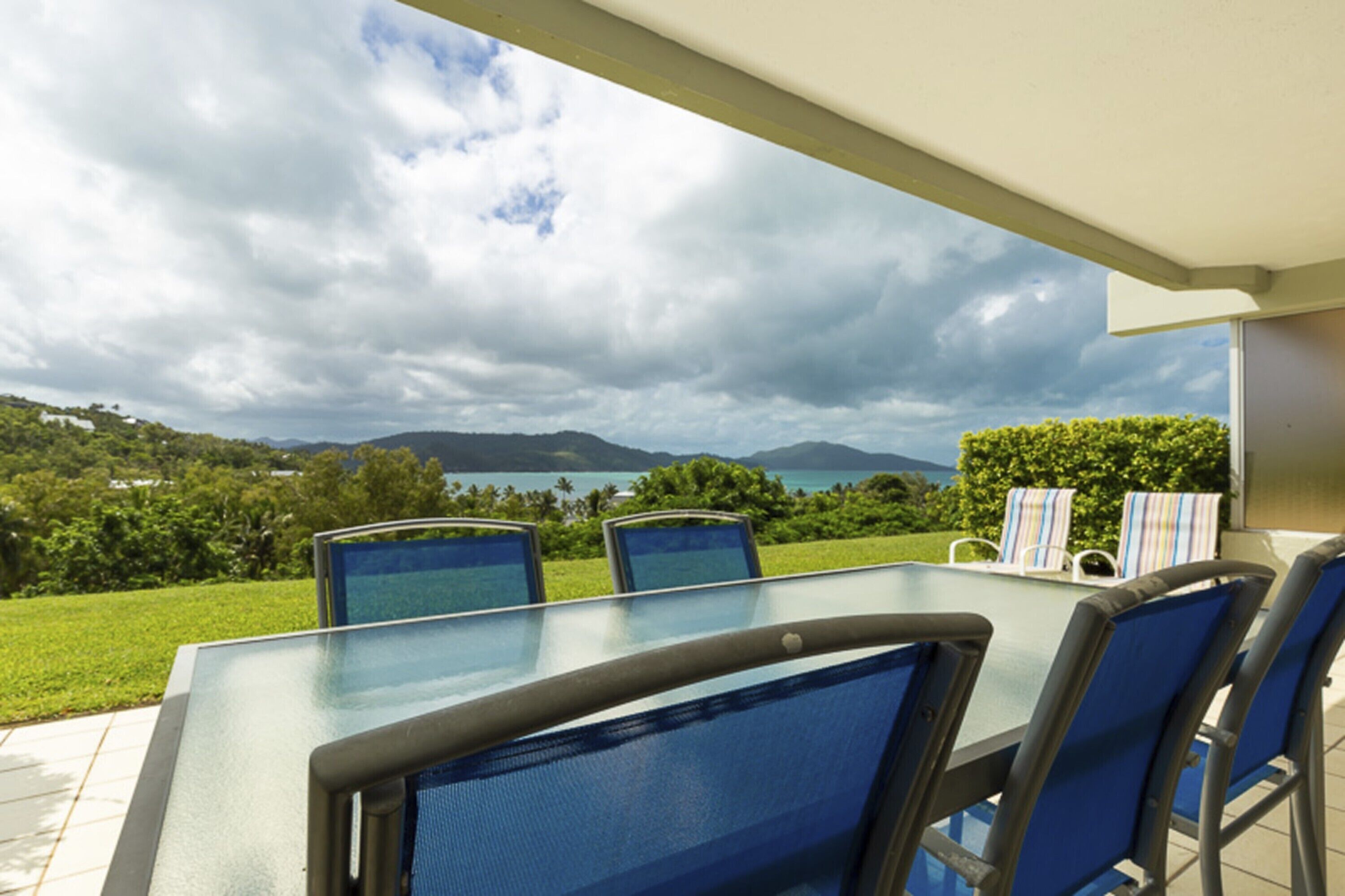 A glass-top table with four blue chairs, a glass balustrade, and a view of a lush landscape with mountains and a body of water.
