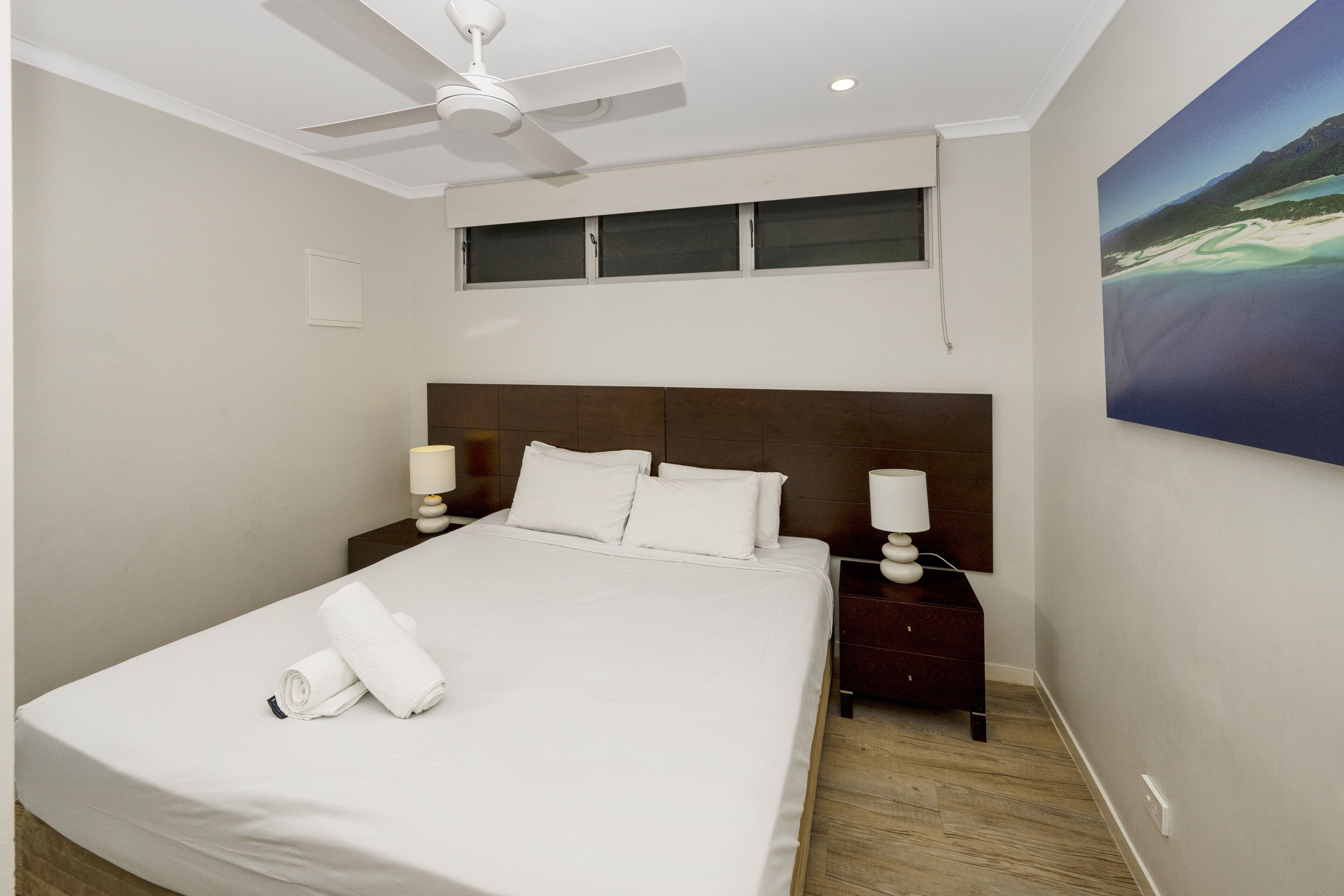 Una cama bien hecha con sábanas y almohadas blancas, un ventilador de techo, una mesita de noche con lámpara, y un cuadro de una playa en la pared.