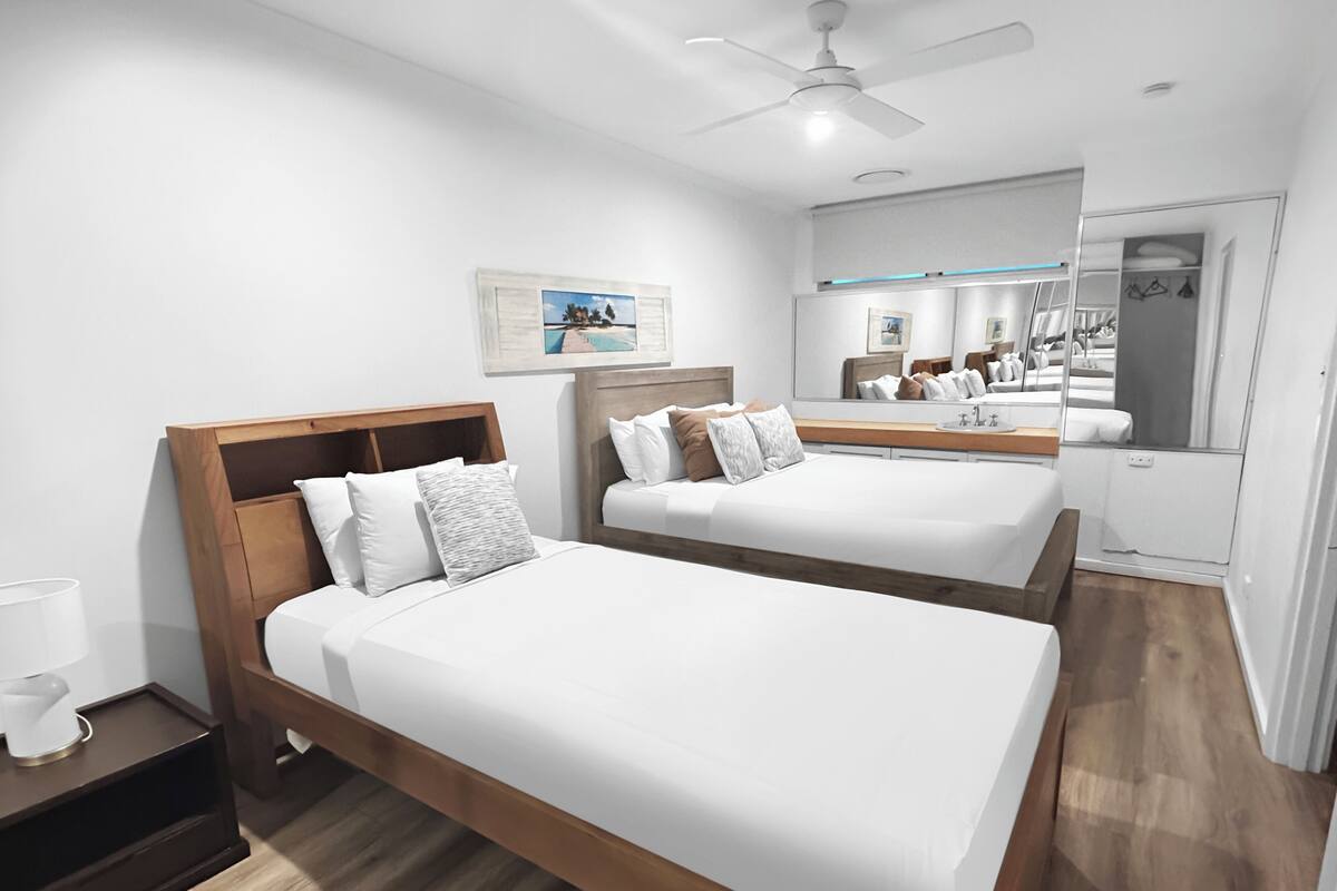 A hotel room with two beds, a ceiling fan, a mirror, and a small bedside table with a lamp.