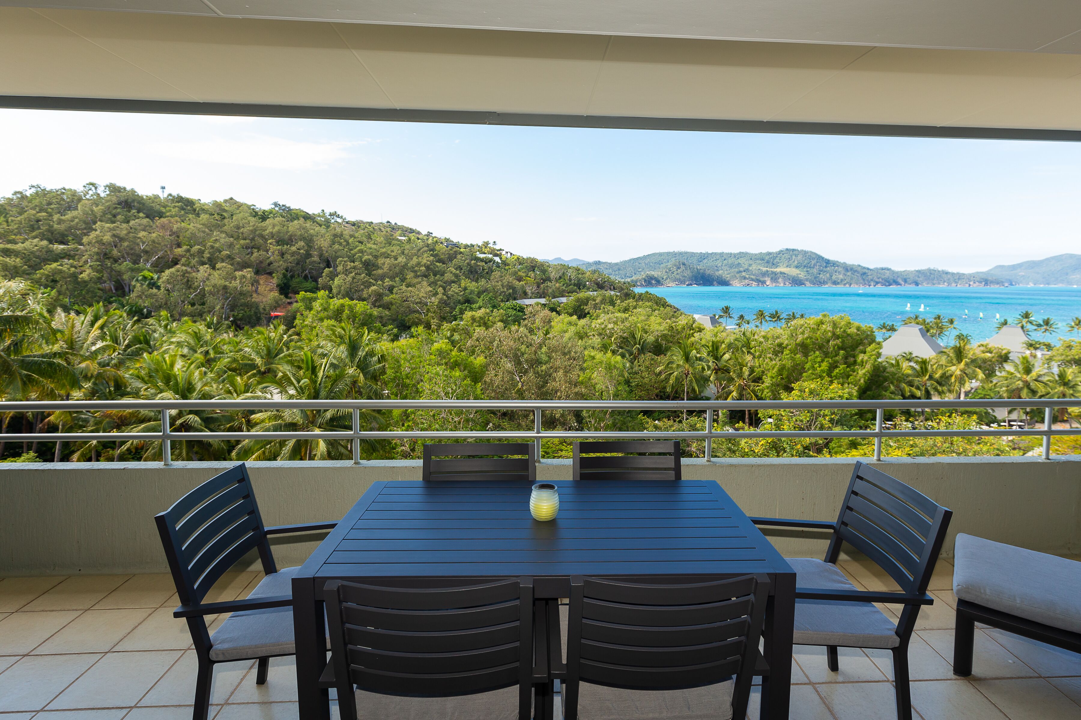 A balcony with a table and chairs, overlooking a coastal landscape with hills and the sea.