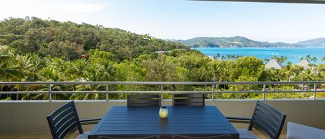 A balcony with a table and chairs, overlooking a coastal landscape with hills and the sea.