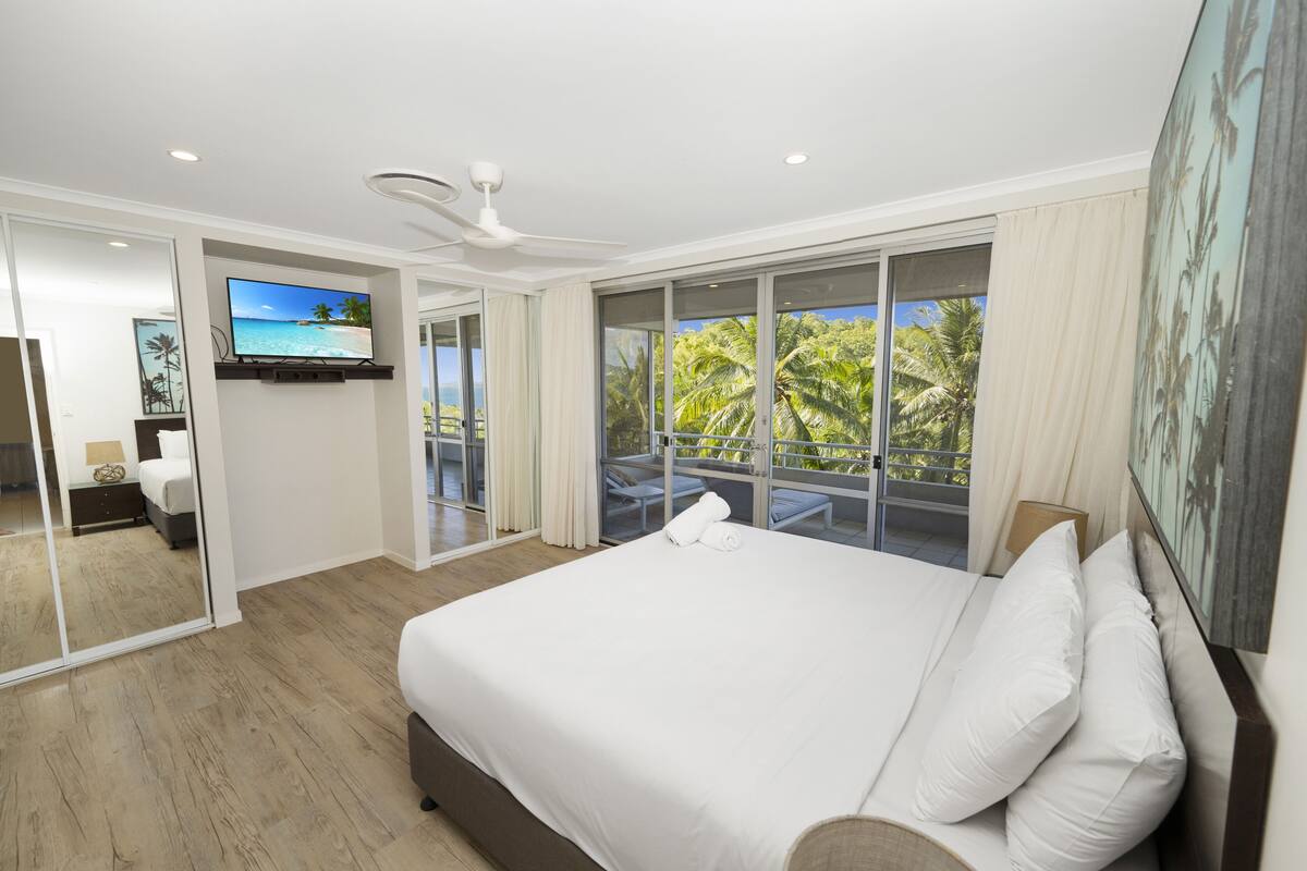 A modern hotel room with a large bed, a ceiling fan, a TV, and a balcony with a view of palm trees.