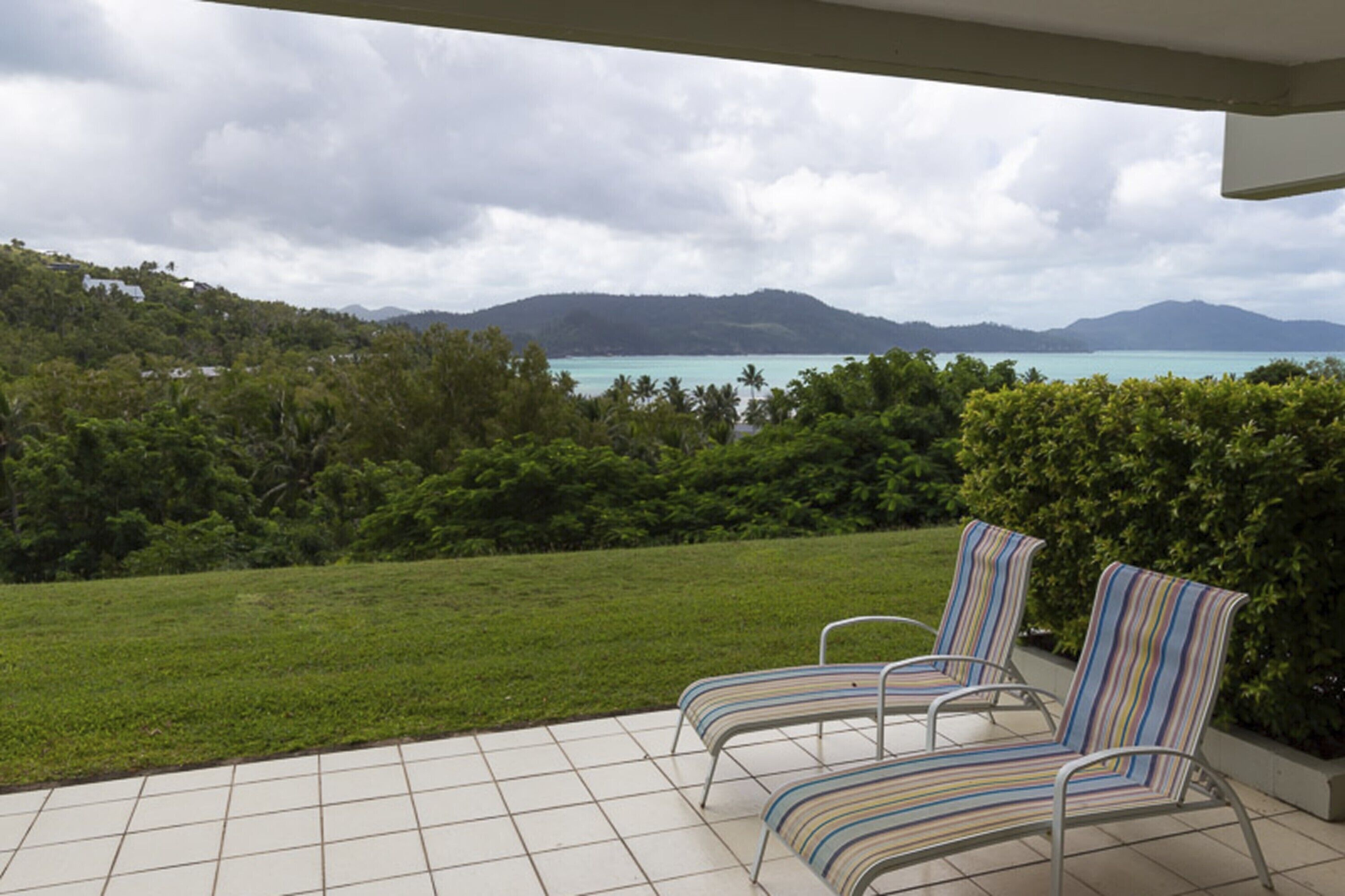 Une terrasse dotée de deux chaises longues à rayures, offrant une vue sur un paysage verdoyant et luxuriant, ainsi que sur la mer et les montagnes lointaines.